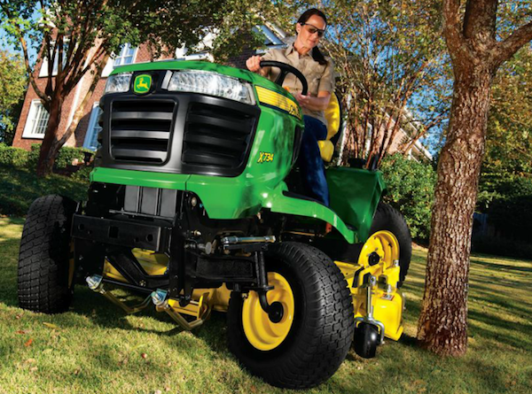 <img src=“X734.png” alt=“Woman with hands on the wheel of a John Deere X734 riding mower next to tree”>