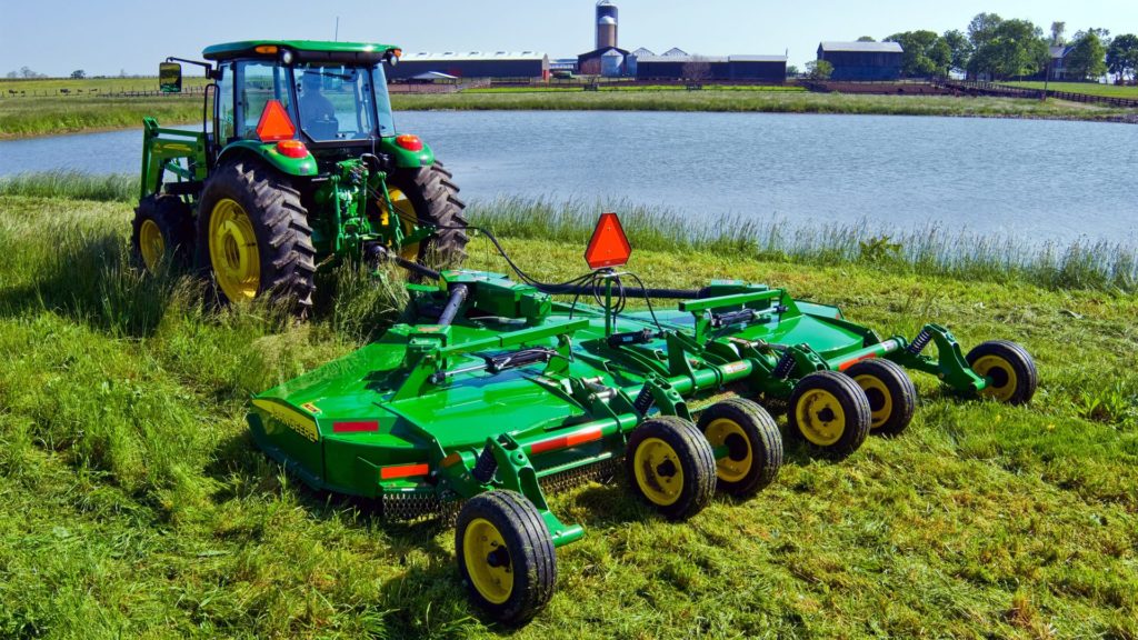 John Deere tractor pulling a rotary cutter to mow grass near a farm pond.