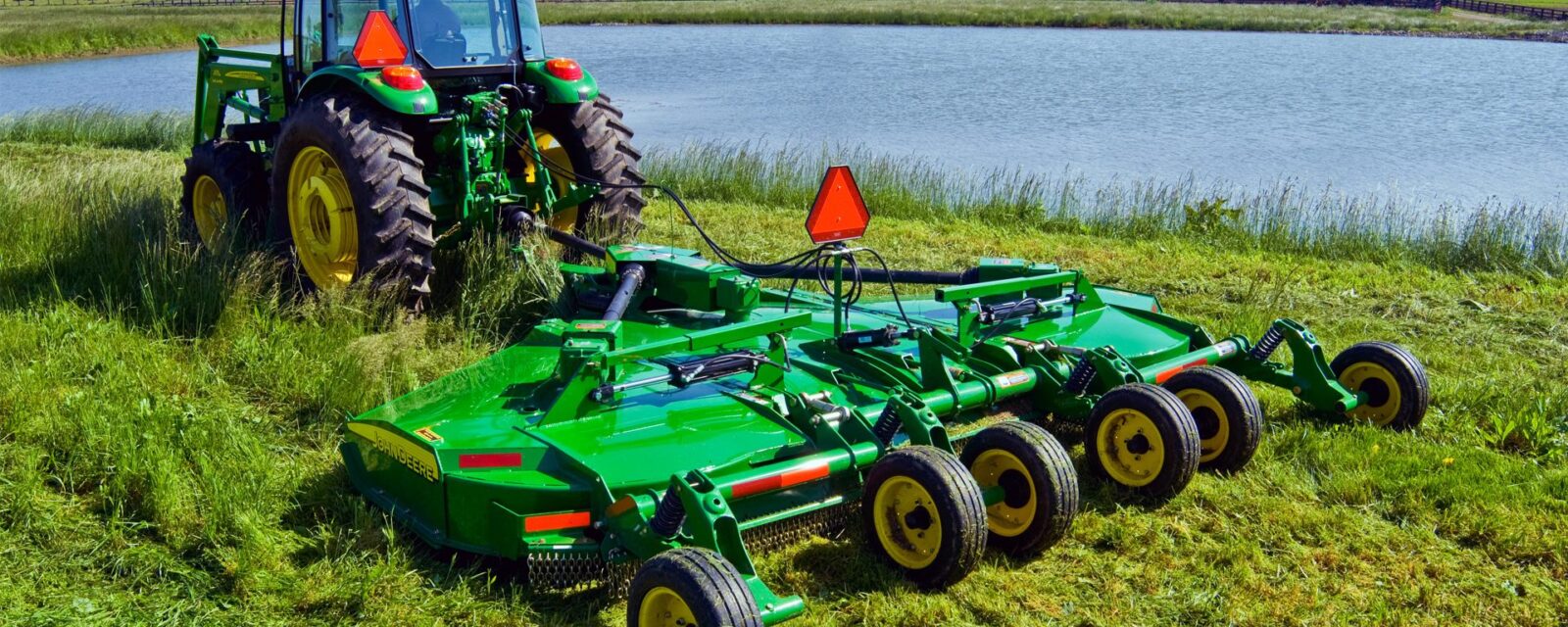 John Deere tractor pulling a rotary cutter to mow grass near a farm pond.