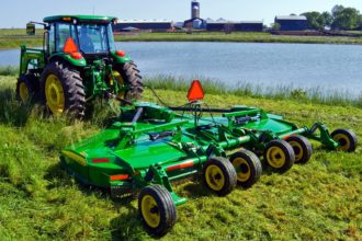 John Deere tractor pulling a rotary cutter to mow grass near a farm pond.