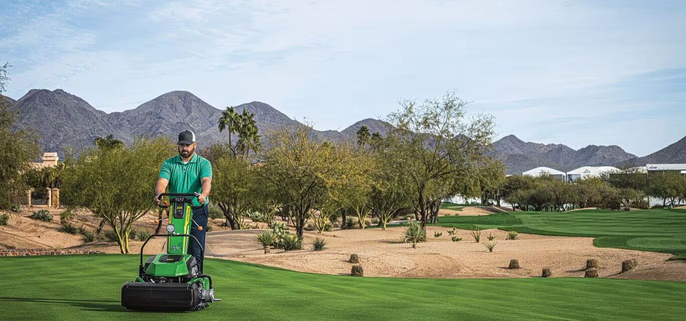 Operator using a John Deere Walking Greens Mower to cut a golf course green with desert landscaping and mountains in the background.