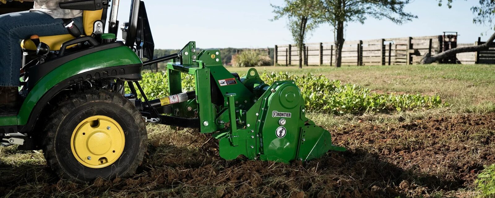 John Deere compact tractor using a Frontier rotary tiller to work soil in a garden.