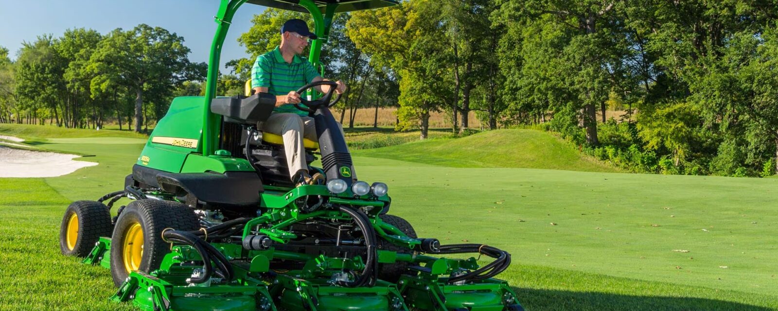 John Deere rough, trim, and surrounds mower cutting turf beside a golf course green.