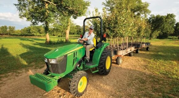 John Deere compact tractor pulling trailers loaded with young trees on a small farm.