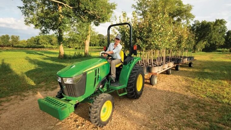 John Deere compact tractor pulling trailers loaded with young trees on a small farm.