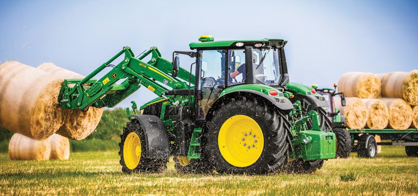 John Deere 6M tractor using a front loader to move large round hay bales in a field.