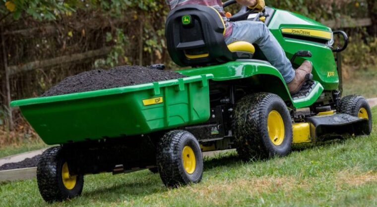John Deere X370 riding lawn mower pulling a green utility cart filled with soil across a grassy lawn.
